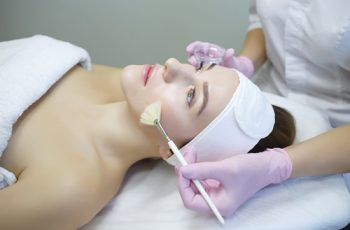 Paciente deitada numa maca, com uma faixa na testa e mão de esteticista passando um pincel com produto no seu rosto, semelhante ao tratamento na clínica Teresa Noviello