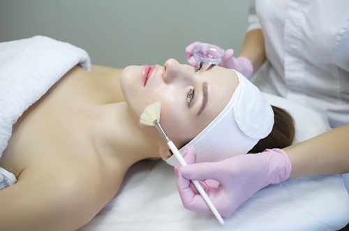 Paciente deitada numa maca, com uma faixa na testa e mão de esteticista passando um pincel com produto no seu rosto, semelhante ao tratamento na clínica Teresa Noviello